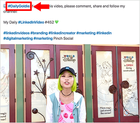 Tämä on kuvakaappaus, joka kuvaa, kuinka Goldie Chan käyttää hashtageja LinkedIn-videopuheluidensa tekstissä. Punaiset huomiotekstit osoittavat tekstissä #DailyGoldie-hashtagin, joka on ainutlaatuinen hänen videopostilleen ja auttaa häntä seuraamaan jakamista. Viesti sisältää myös muita asiaankuuluvia hashtageja, jotka auttavat ihmisiä löytämään hänen videonsa, mukaan lukien #LinkedInVideo. Videokuvassa Goldie seisoo joidenkin ovien edessä World of Disney -näyttelyssä. Hän on aasialainen nainen, jolla on vihreät hiukset. Hänellä on yllään musta LinkedIn-lippis, musta choker-kaulakoru, vaaleanpunainen macaron-painettu paita ja sininen ja valkoinen takki.
